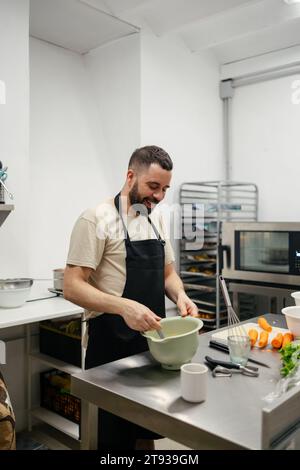 Young bearded chef dressed in a kitchen apron smiling and stirring with a spoon in a well-equipped kitchen Stock Photo