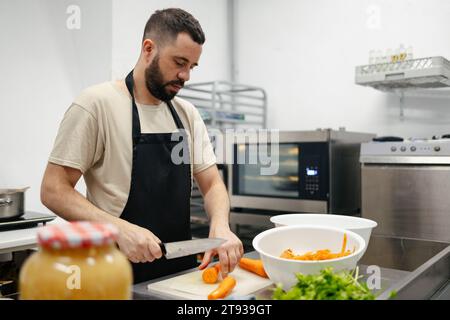 Young bearded chef dressed in a kitchen apron cutting carrots in the table of a well-equipped kitchen Stock Photo