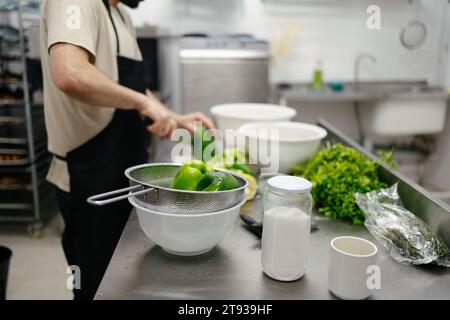 Close-up of green peppers while in the background there is an out-of-focus and unrecognizable chef cutting peppers in a professional kitchen Stock Photo