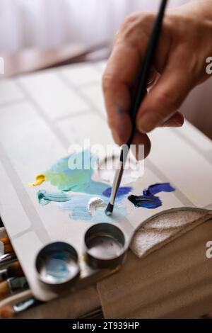 Close-up of the unrecognizable hand of a painter picking up paint with a paint brush from a paint palette with different brushes around it Stock Photo