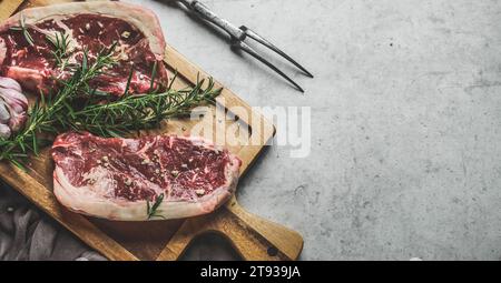 Raw beef steaks background with rosemary and green pepper on wooden cutting board with butcher fork. Close up. Preparing fresh meat. Top view with cop Stock Photo