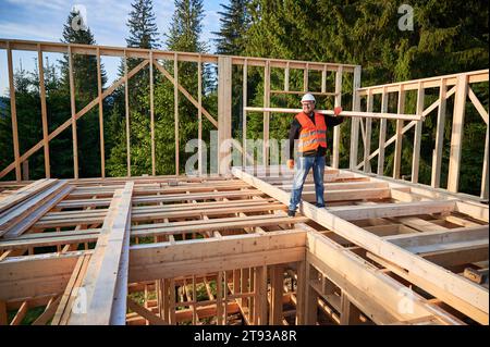 Carpenter builds wooden frame house near woods. Man holds large beam on shoulder, wearing work clothes and helmet. Concept of modern and sustainable building. Stock Photo