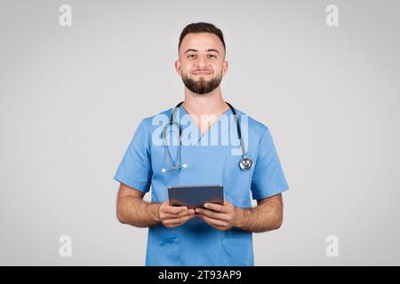Smiling male doctor with tablet, blue coat, grey background Stock Photo