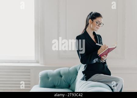 Focused professional woman writing in notebook, seated on a stylish blue couch in a bright room Stock Photo