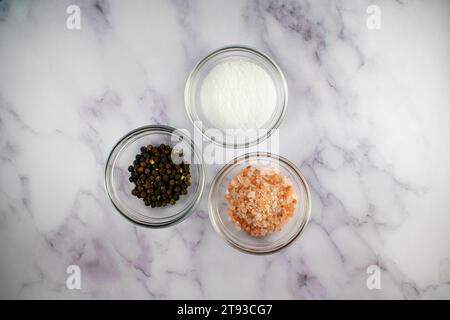 Top down flat lay photo showing salt, Himalayan pink salt, and black peppercorns on a marble backdrop Stock Photo