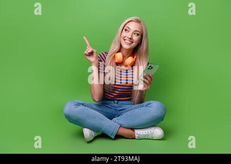 Full body photo of girl new youtube premium service download music point finger copyspace headphones ad isolated on green color background Stock Photo