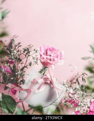 White greeting card with ribbon and rose flowers at pink background with green leaves frame. Top view with copy space. Stock Photo