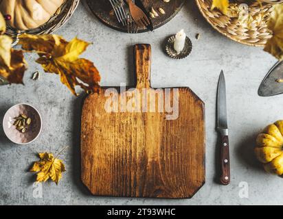 Food background with empty wooden cutting board, pumpkin, knife, autumn leaves and utensils at grey table. Cooking at home with seasonal ingredients. Stock Photo