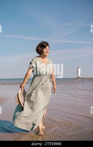 A woman in a flowing dress stands on a sandy beach, from the sun with a ...