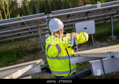 Female technician using a cordless drill on solar panel structure in a field. Alternative energy ecological concept image. Stock Photo