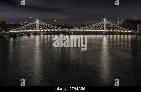 London, UK - Nov 20, 2023 - Nocturnal view of floodlit Albert Bridge from Chelsea Bridge with the reflection of the light in very calm water of River Stock Photo