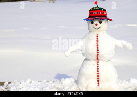 A funny snowman in red hat stands on snow covered meadow in winter, Christmas, New Year holidays, children entertainment and sports, outdoor Stock Photo