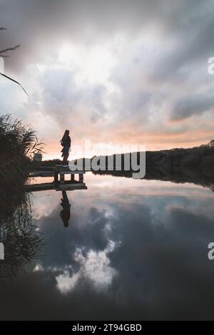 Lady aged 20-25 standing on a wooden pier by a pond watching the sunset over the town of Oostende, western Belgium. Dramatic red-orange sky. Stock Photo