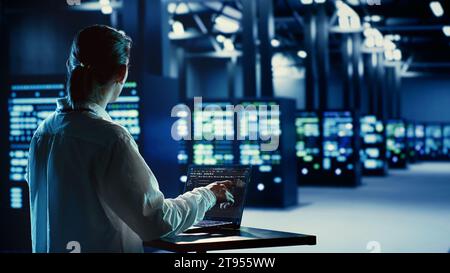 Asian system administrator running code and programming on laptop, troubleshooting and upgrading server farm equipment designed to accommodate mainframes, networking racks and storage arrays Stock Photo