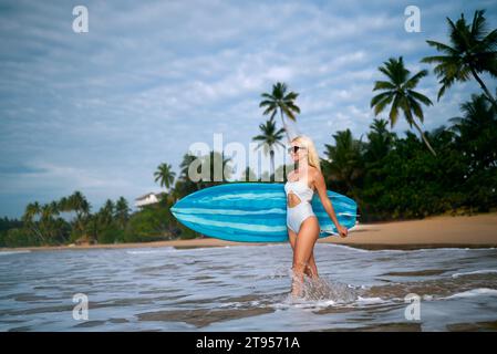 Blond woman in white bikini carries blue surfboard on tropical beach, palm trees in background, leisure activity in summer, ocean waves and sandy Stock Photo