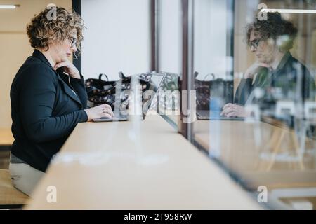 Middle-aged businesswoman sitting at working desk next to the window working on lap top. Side view shot. Reflection from window. Stock Photo