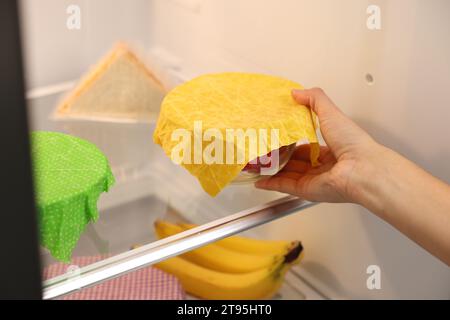 Woman putting bowl covered with beeswax food wrap into refrigerator, closeup Stock Photo