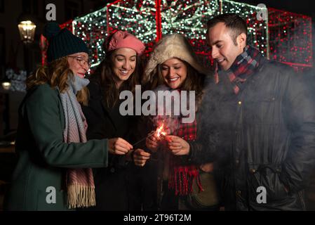 Happy young Hispanic male and female friends looking down while standing on street and burning sparklers together against glowing garland during night Stock Photo