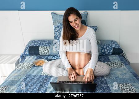 From above of smiling pregnant female speaking on video call on netbook while sitting on comfortable bed at home Stock Photo