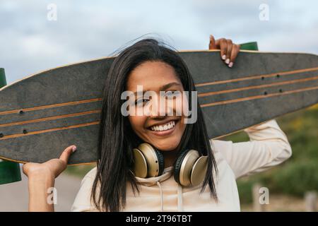 Young smiling ethnic cheerful female standing with longboard behind head and headphones on neck looking at camera Stock Photo