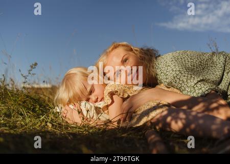 Smiling woman wearing green summer dress with flower pattern lying on meadow and embracing little blond haired daughter while enjoying sunny weekend Stock Photo
