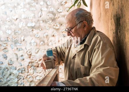 Side view of focused senior male carpenter wearing casuals and glasses shaping wooden plank with plane tool while standing by wall Stock Photo