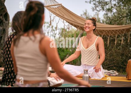 Positive smiling female yoga teacher with closed eyes sitting in Padmasana pose on mat in front of group of women during meditation session in nature Stock Photo