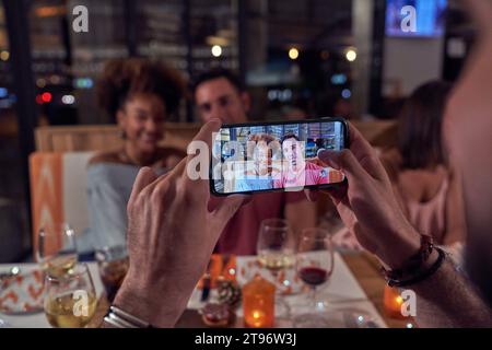 Unrecognizable man taking photo of young smiling friends sitting at table in modern restaurant while enjoying beverages in evening Stock Photo