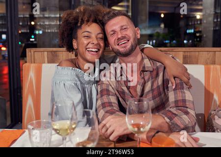 Delighted black woman and man hugging and looking at camera with smile while sitting together at table with alcohol drinks in restaurant Stock Photo