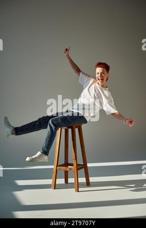 overjoyed queer person having fun while sitting on stool and showing thumbs up in sunlight on grey Stock Photo