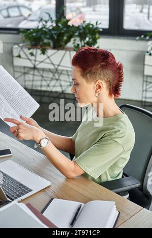 side view of stylish non-binary person working with documents near laptop and mobile phone in office Stock Photo