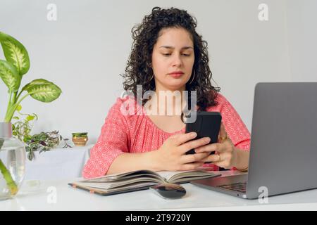 young Latin Venezuelan woman with curlers, home worker sitting in office with her laptop concentrated sending messages on phone, copy space. Stock Photo
