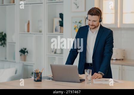 Focused businessman in a suit working remotely with a headset, showcasing productivity and professionalism in a well-lit home office Stock Photo