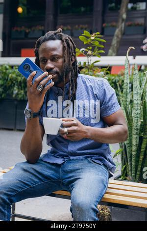 Vertical image of African man with dreadlocks, happy sitting outdoors sending voice message on his phone, communicating with his girlfriend while rest Stock Photo