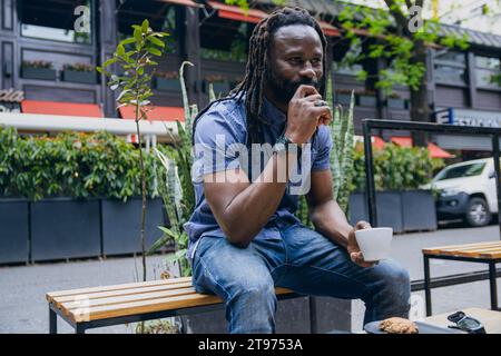 young african man with dreadlocks sitting outdoors drinking coffee and thinking with his hand on his mouth, looking ahead meditating and enjoying rest Stock Photo