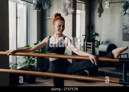 Redhead fitness enthusiast perfecting ballet stretches at the barre in a modern urban gym Stock Photo