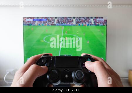 A male hand holding a play station 4 controller with football video game in a smart tv at background Stock Photo