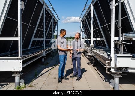 Two employees with digital tablet talking on rooftop beside refrigeration installation Stock Photo