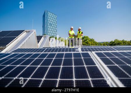 Two technicians utilizing a tablet PC while inspecting solar panels on the rooftop of a corporate building Stock Photo
