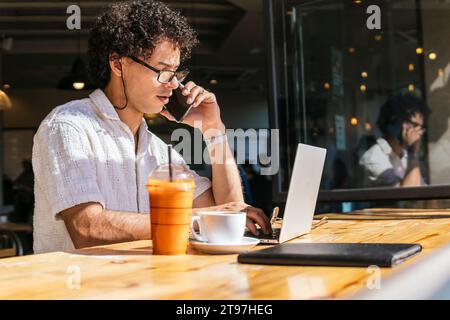 Young freelancer talking on smart phone and using laptop in cafe Stock Photo
