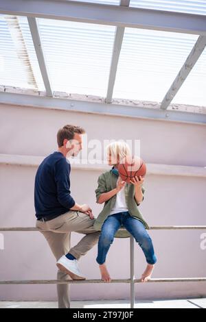Father talking to son holding basketball and sitting on railing Stock Photo