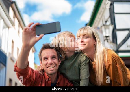 Smiling father taking selfie with son kissing mother on sunny day Stock Photo