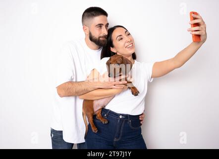Young couple with her dachshund dog taking a selfie standing isolated over white background. Stock Photo