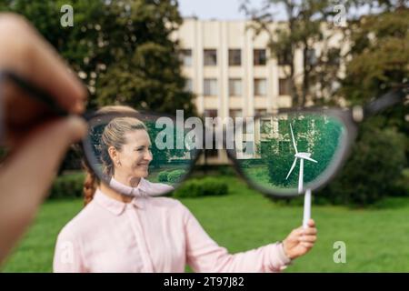 Smiling businesswoman holding wind turbine model seen through concave eyeglasses at park Stock Photo