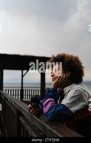 Thoughtful young woman standing with camera near railing Stock Photo
