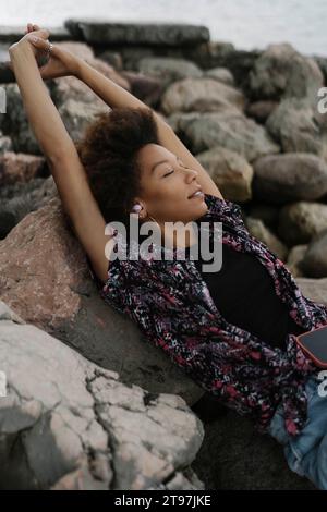 Young woman with eyes closed and arms raised relaxing on rocks Stock Photo