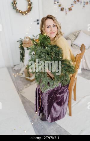 Beautiful woman sitting on chair and holding bouquet of pine tree branches at home Stock Photo