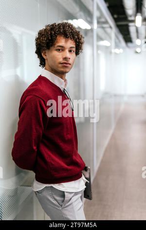 Businessman with hands behind back holding laptop and standing in front of glass at office Stock Photo