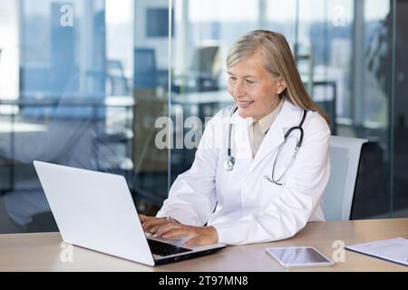Senior smiling gray-haired female doctor consults, works and types on laptop online. Sitting at the desk in the office of the clinic. Stock Photo