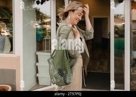 Smiling woman with hand in hair holding mesh bag by eco-shop Stock Photo